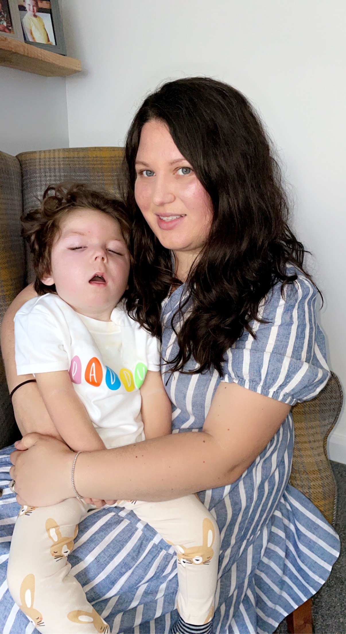 A woman with dark hair holds a young child in her arms. The child wears a colorful "Daddy" shirt and rests peacefully.