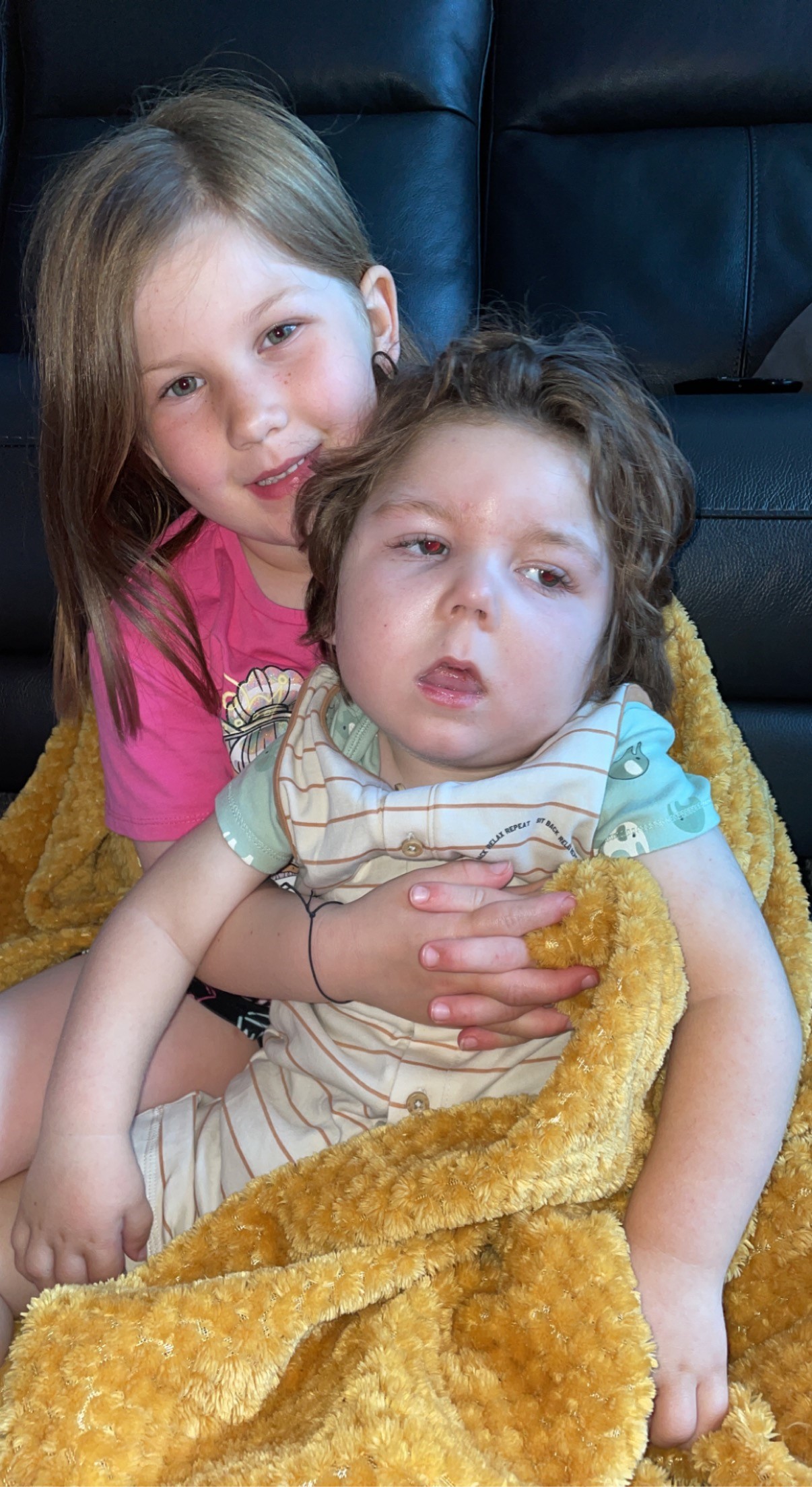 A young girl with light brown hair and a pink shirt holds a younger child with curly hair, wrapped in a yellow blanket, on a couch